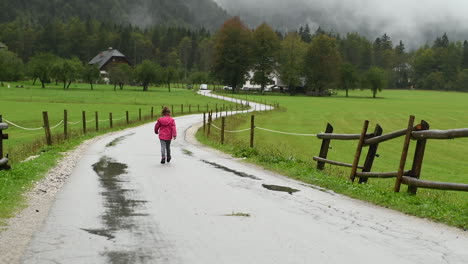 little girl playfully walking in rain on country road, farmhouse in background, alpine valley, zoom in, from behind facing away