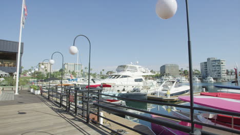 pov of a person walking at boardwalk along newport beach harbor with yachts and boats on a sunny summer day