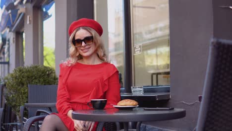 young woman smiles and adjusts stylish sunglasses. woman in a red dress and beret sitting with a cup of coffee and a croissant on a table on the street