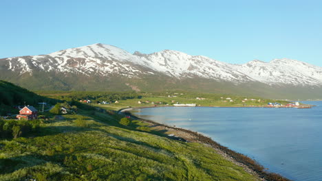 aerial low, drone shot over houses in a small village on the coast of the barents sea, snowy mountains in the background, sunny day, in troms, nordland, north norway