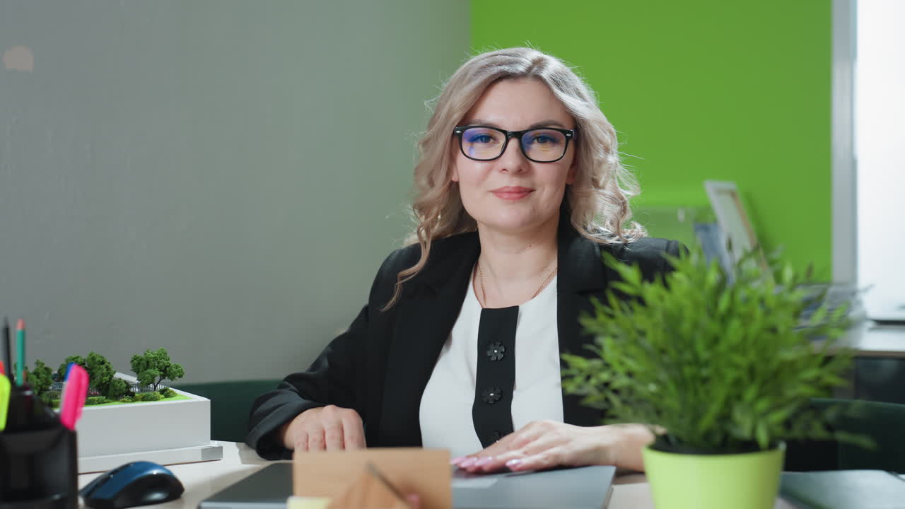 business mogul with blonde wavy hair wearing glasses confidently closes laptop while seated at modern office desk with building model, green plant, pen holder, and stylish minimal decor
