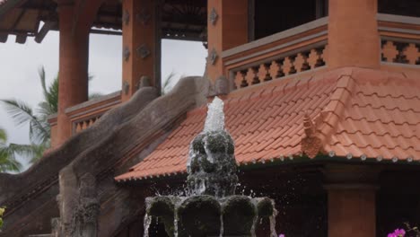 traditional stone fountain with cascading water, surrounded by vibrant flowers, stands in front of traditional balinese architecture