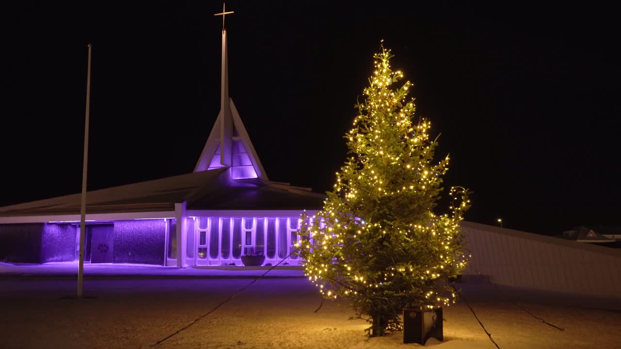 강한 겨울 폭풍의 안정적인 은 크리스마스 불빛으로 인 크리스마스 트리를 불어넣고, 배경에는 보라색으로 조명된 교회가 있습니다.