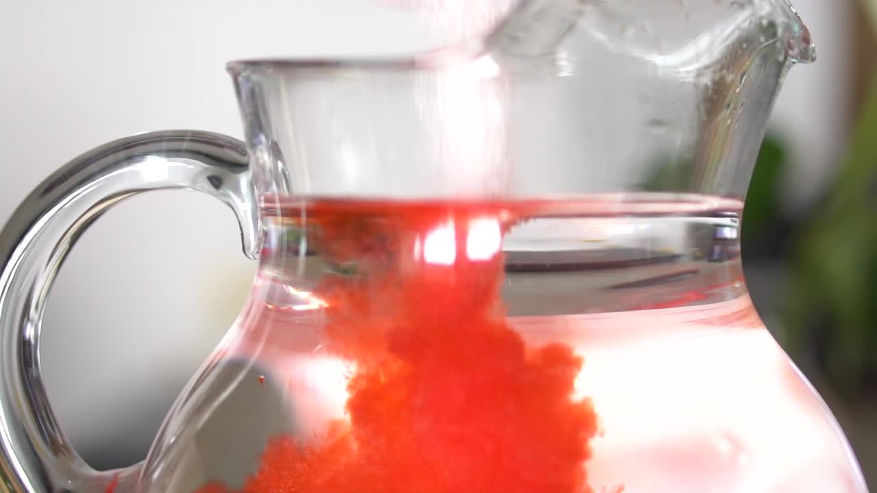 Red powder drink mix pouring into glass pitcher of water.