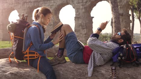 happy young couple backpackers tourists sitting lying on a log trunk reading book using smartphone in front of ancient roman aqueduct ruins in parco degli acquedotti park in rome at sunrise romantic with sleeping bag slow motion