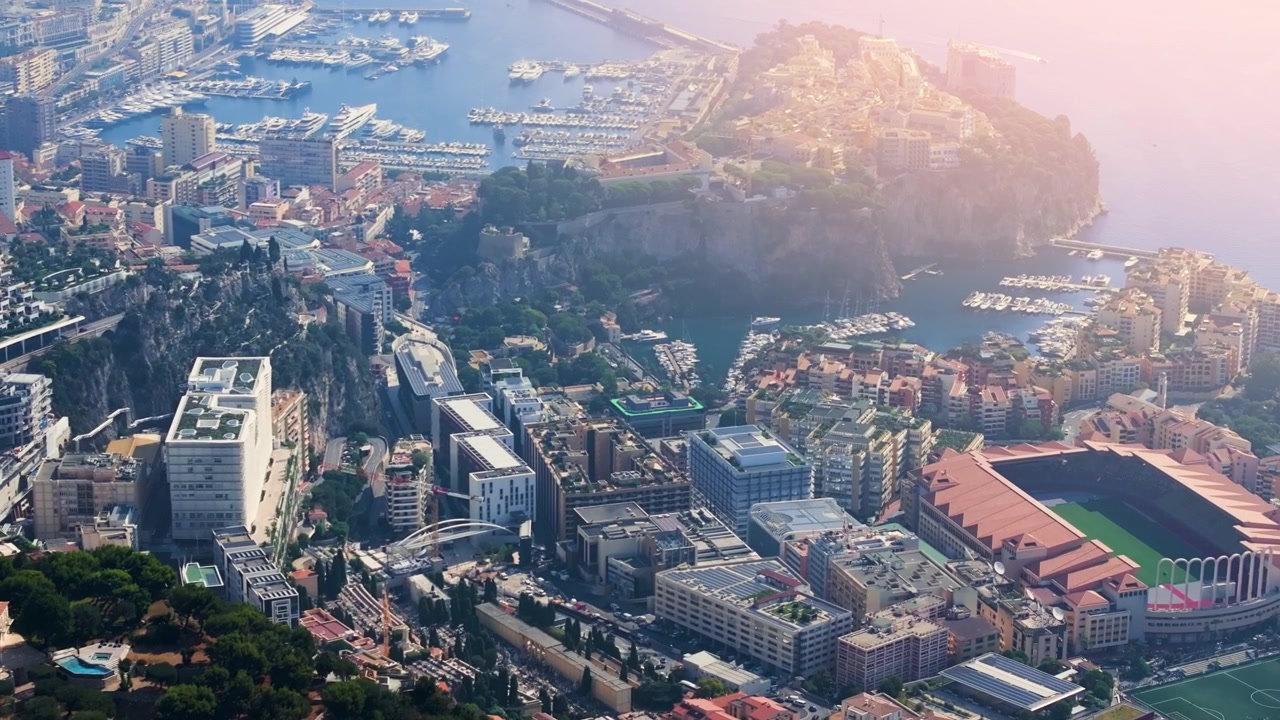 Monaco è mostrata dall'alto con un focus sul porto e sugli edifici vicini. La vista cattura la disposizione della città, così come colline e acqua sullo sfondo durante il giorno