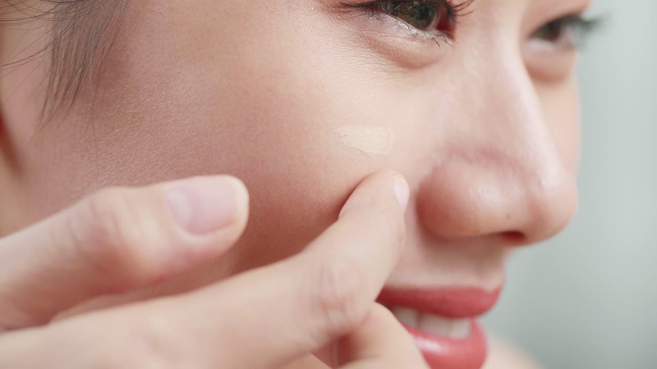 美しい若いアジア人の女性が顔にクリームを塗り自宅でメイクをしている間に笑顔を浮かべています