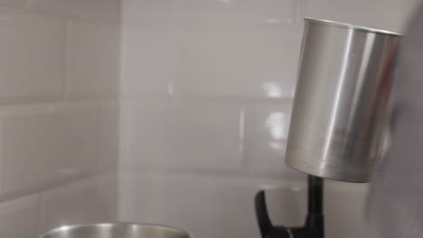lady removes metal jug from steaming pot in kitchen, cleans bottom, carefully holds glass cup with wick to pour melted liquid from jug, candle making against tiled wall background