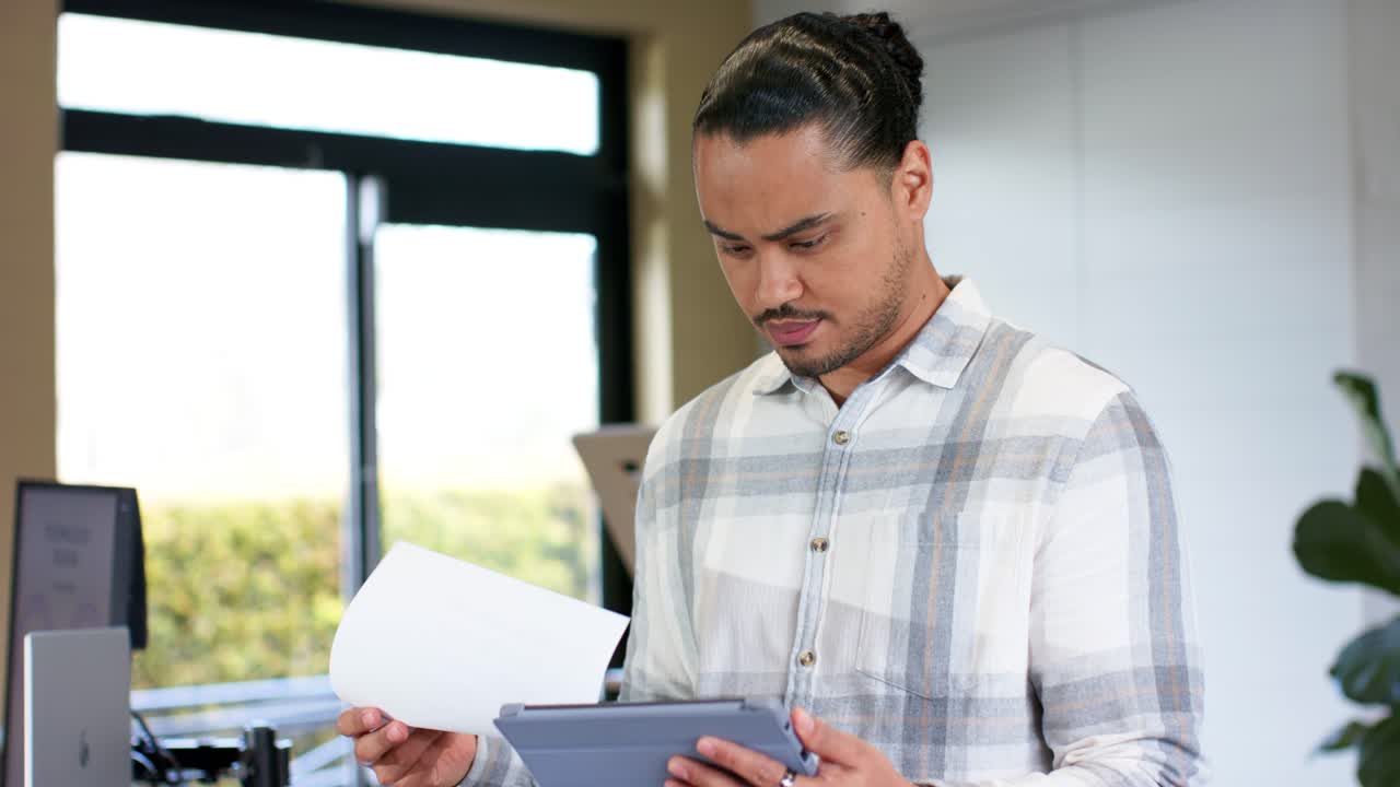 Mid adult man studying tablet at office standing desk by window retrieving paper, comparing info
