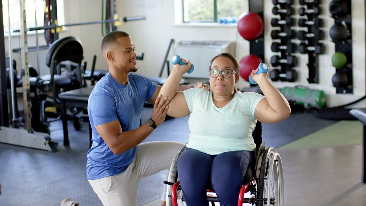 Physical therapist assisting woman with paraplegia in wheelchair lifting dumbbells in gym