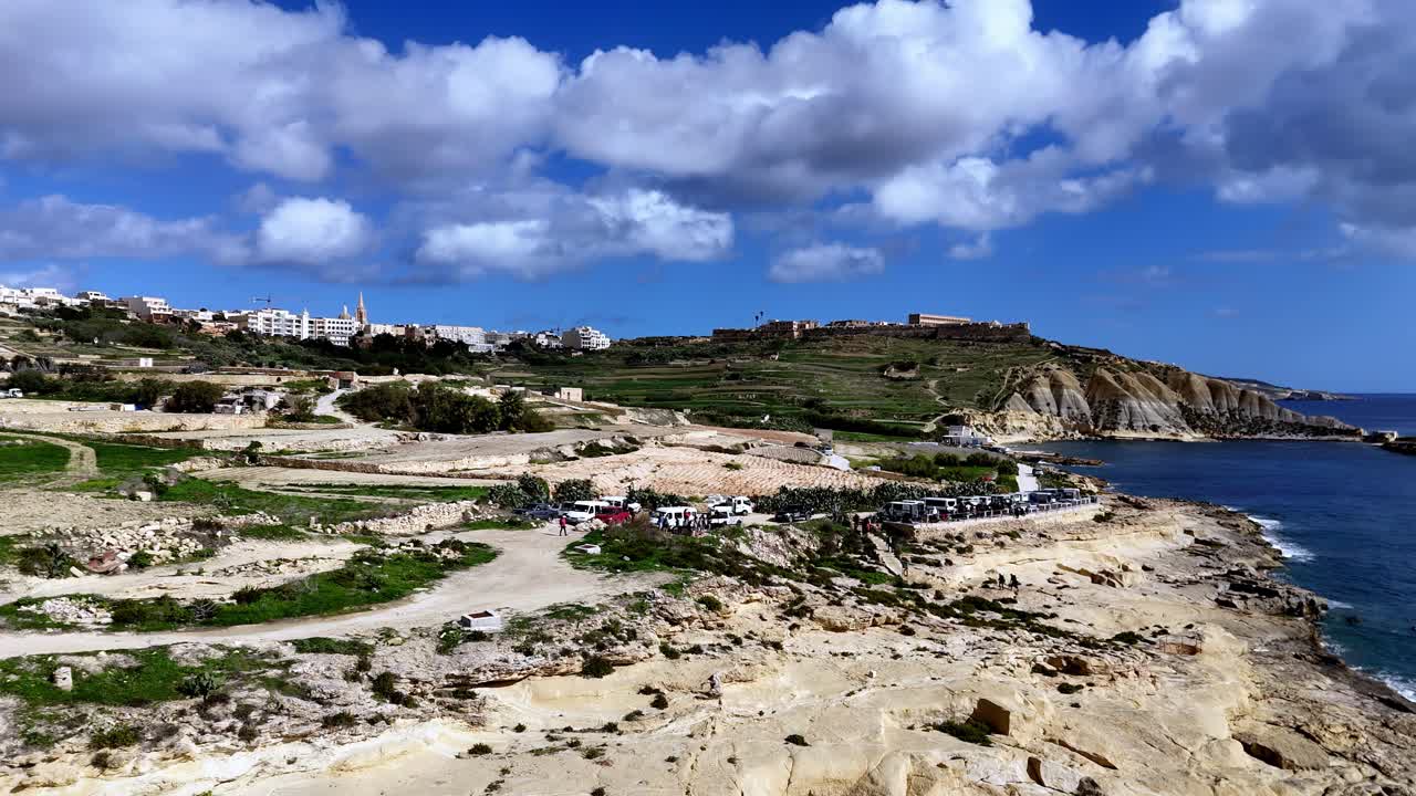 Panorama mozzafiato dall'alto della costa di Gozo a Malta, durante una soleggiata giornata primaverile, con scogliere di calcare dorato e un mare Mediterraneo blu intenso sotto soffici nuvole bianche, vicino all'isola di Comino.