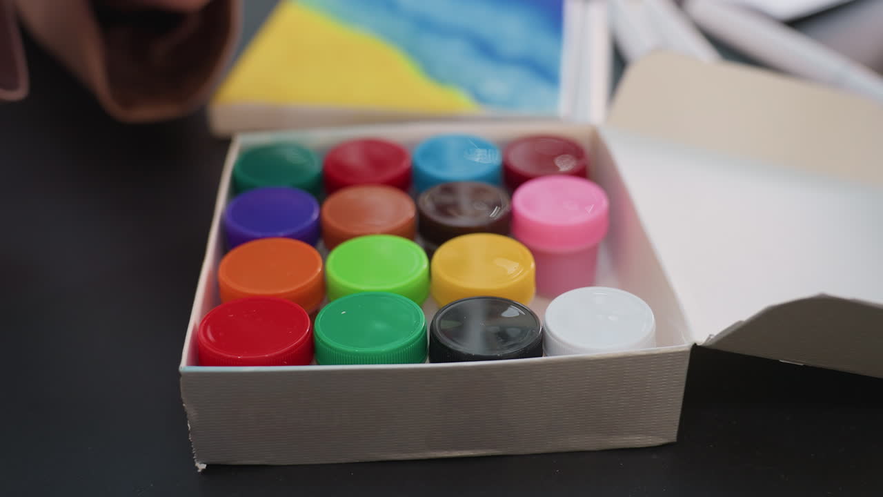 여성 아티스트가 다가오는 프로젝트를 위해 신중하게 다채로운 페인트 통을 고르고 있어요. 아티스트는 페인팅 세션을 준비하기 위해 다양한 색조의 페인트 용기를 꼼꼼하게 선택하고 분류하고 있습니다.