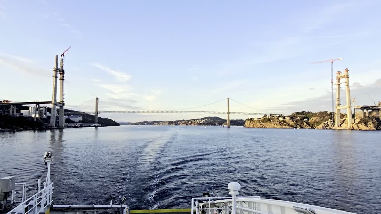 Ship sails south through fjord showing new Sotra Bridge towers and old bridge in sunset light