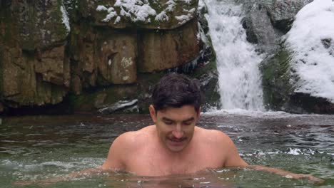 Man-gets-out-of-the-cold-water-by-a-beautiful-waterfall-in-snowy-mountains-during-winter-day---static-shot,-slow-motion