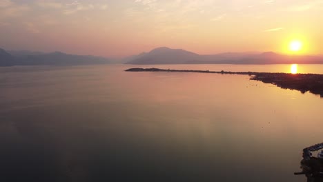 Toma-Aérea-Del-Lago-Garda-Al-Amanecer-Con-Sirmione-Y-Montañas-En-La-Parte-De-Atrás