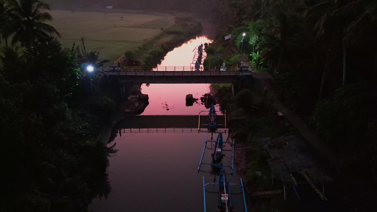 Early morning drone footage reveals a bridge crossing a quiet river, glowing in a golden hue. Warm tones reflect off the water, creating a magical tropical landscape from above