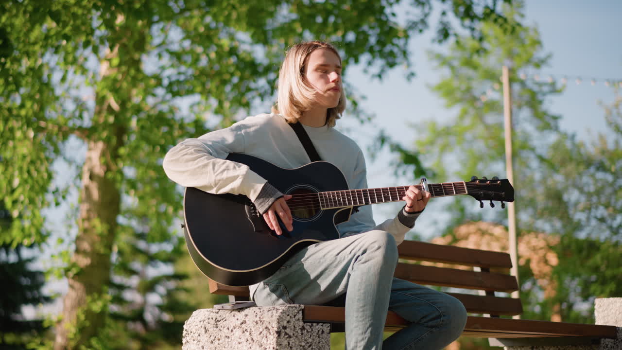 giovane chitarrista che suona la chitarra acustica all'aperto sotto alberi illuminati dal sole, posa rilassata su una panchina di pietra, maglione e jeans, delicato strumming e melodia emotiva, atmosfera di cinguettio di uccelli, profondità cinematografica