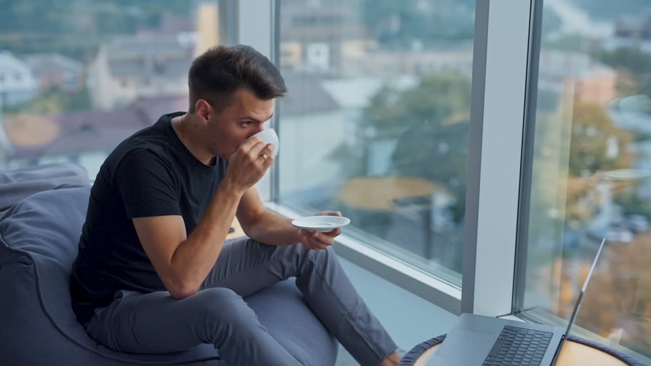 Brunet young man drinking coffee at lunch break sitting near panoramic window. Thoughtful male sitting in front of laptop. Blurred backdrop.