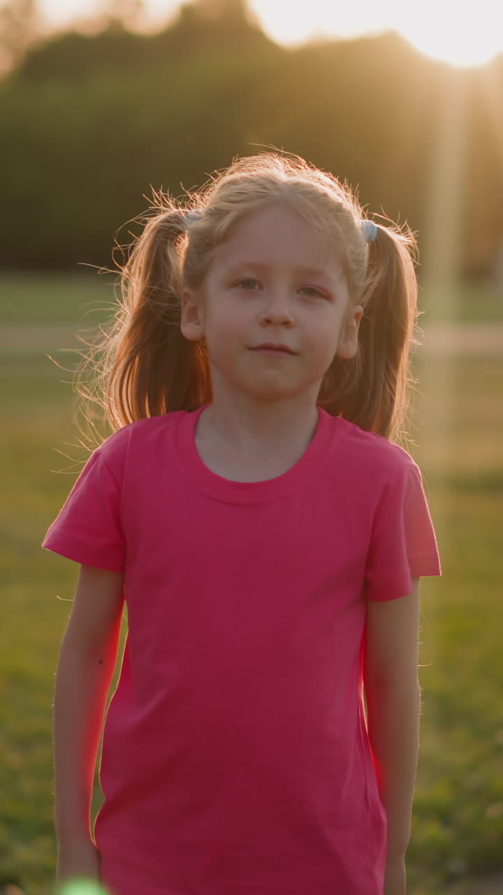 Pretty little girl shakes ponytails standing on park meadow at sunset slow motion. Long haired child has fun at countryside. Leisure and game