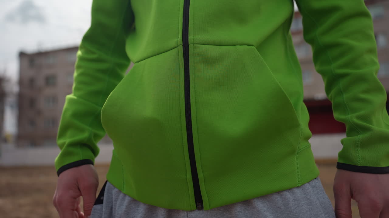 Persona discreta con las manos en los bolsillos en el exterior, instantánea serena de alguien con sudadera cerca de una zona residencial, escena tranquila de una persona con sudadera verde en un barrio apacible