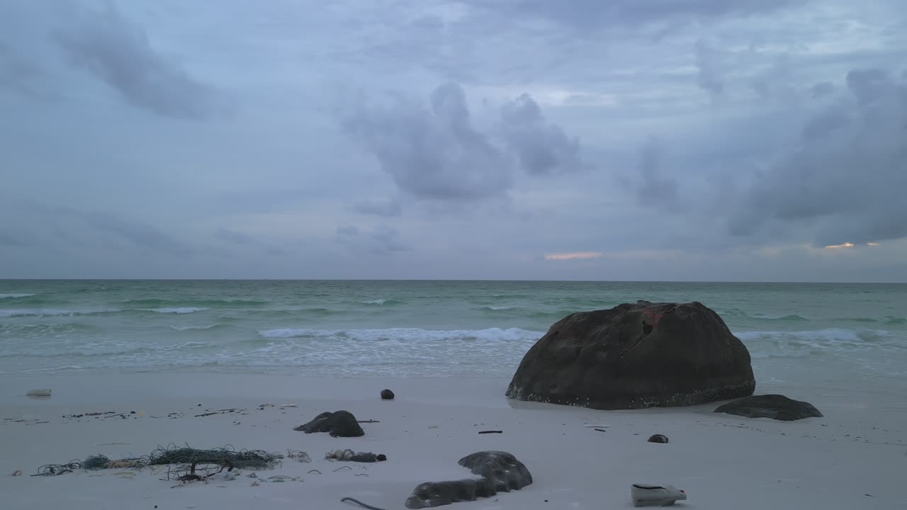 Orbital drone movement around a large rock on a white sandy beach, with turquoise ocean in the background.