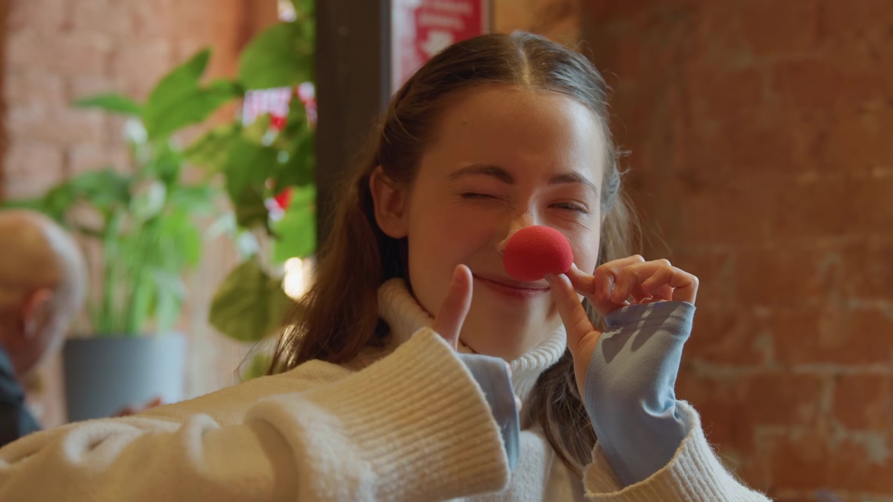 Close up of young woman holding and examining small red object in her hand. scene is set indoors with soft lighting featuring blurred green plants and warm toned brick walls as background