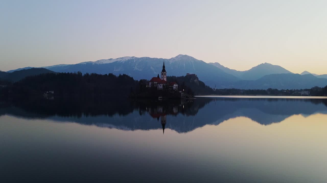 Drone glides toward Bled's island church, revealing dawn's colors behind peaks