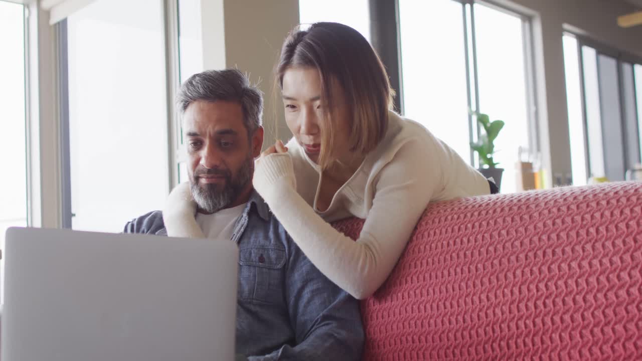 una pareja feliz y diversa sentada en la sala de estar usando una computadora portátil y hablando