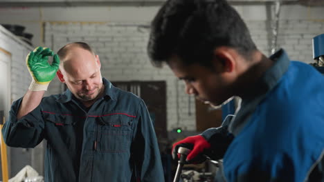 engineer in mechanical workshop working on car engine, tightening nut with red gloves while colleague observes and wipes head, background features work tools, bright light