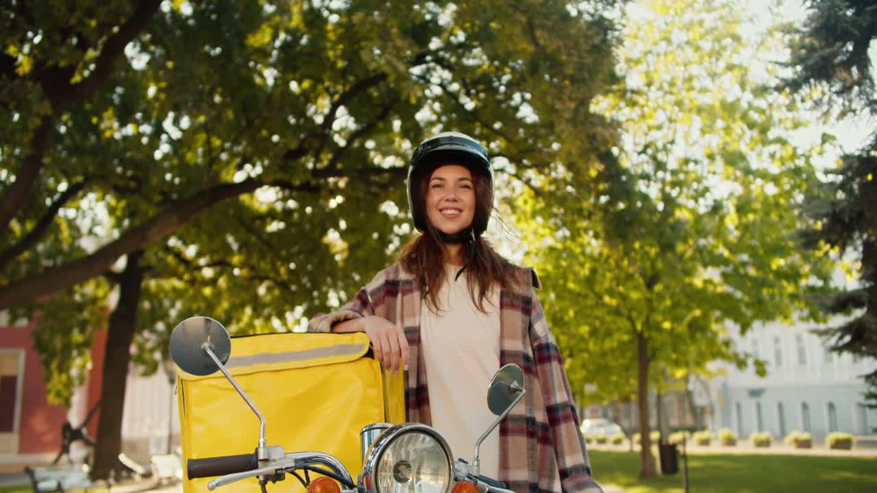 Portrait of a happy brunette courier girl in a checkered shirt wearing a White motorcycle helmet with a yellow bag for food delivery on the street in summer