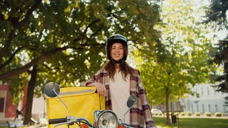 Portrait-of-a-happy-brunette-courier-girl-in-a-checkered-shirt-wearing-a-White-motorcycle-helmet-with-a-yellow-bag-for-food-delivery-on-the-street-in-summer