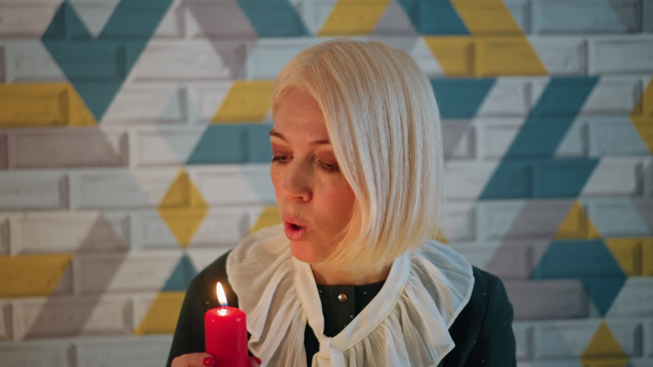 Kaukasisk kvinde holder rødt lys i intime juleatelieromgivelser med geometrisk flisevæg, vintage flæsebluse, blødt varmt lys, eftertænksomt udtryk, puster til en lille flamme, hyggelig højtid.