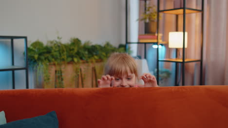 Child-girl-playing-hide-and-seek-peekaboo-game-near-sofa-at-home-alone-looking-at-camera-and-smiling