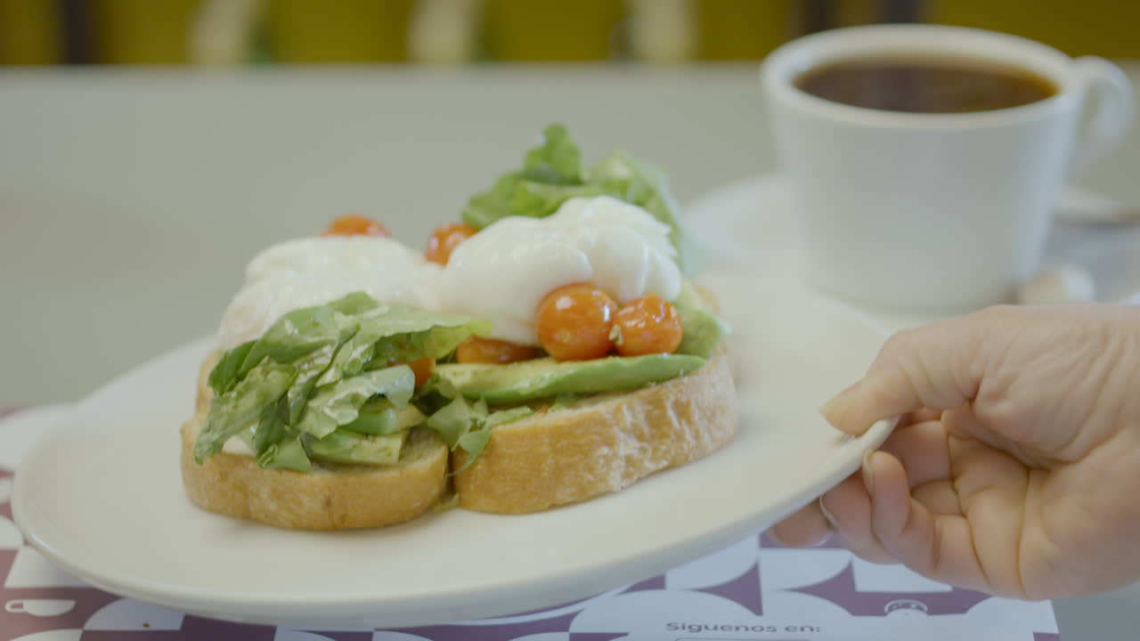 Close-up of a plate with rustic bread toast topped with avocado, lettuce, cherry tomatoes and poached eggs, next to a cup of hot coffee