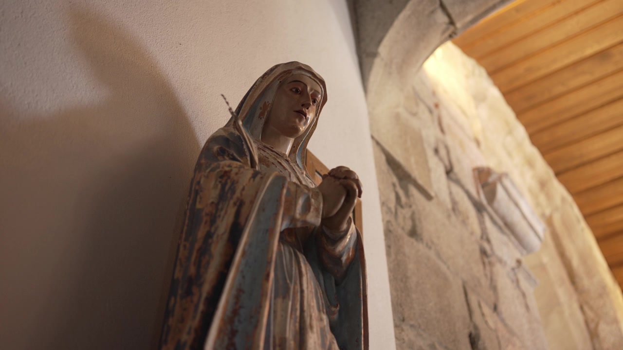 Statua consumata dalle intemperie di una figura in preghiera in un ambiente rustico, che proietta un'ombra contro un muro di pietra