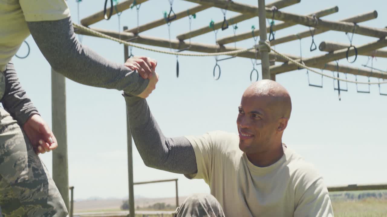 Female soldier helping diverse male colleague with injured leg at army obstacle course in the sun