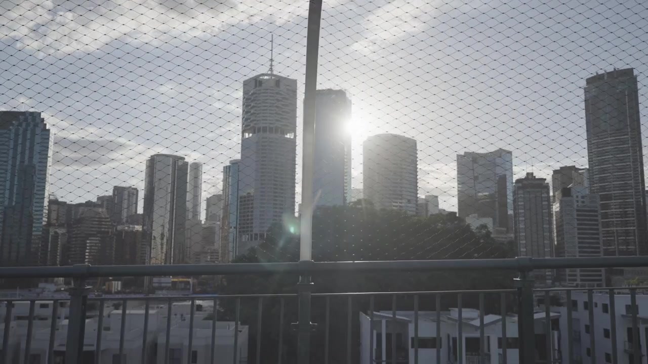 Raios de sol atravessam uma cerca de arame com o horizonte da cidade de Brisbane visível ao fundo em um mirante à beira-rio
