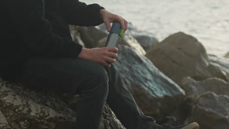 man-unscrews-thermos-and-pours-hot-drink-into-cup-as-sea-waves-wash-over-rocks-in-background-during-golden-hour