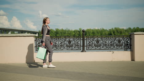 lady in grey clothing walking near riverfront with a shopping bag in hand, passing by a metal fence, background features blur of a bridge, greenery, and clear sky on a sunny day