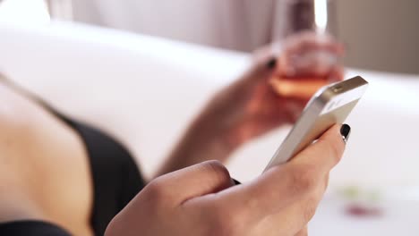 beautiful woman relaxing in milk bath lying in bathtub and drinking sparkling wine, using smartphone browsing social media