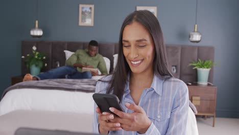 Vídeo-De-Una-Pareja-Diversa-En-El-Dormitorio,-La-Mujer-Sonriendo-Usando-Un-Teléfono-Inteligente-Sentada-En-Primer-Plano