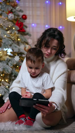 attractive caucasian brunette woman holds a cute baby boy in hands. mother shows a smartphone. christmas tree at backdrop. vertical video.