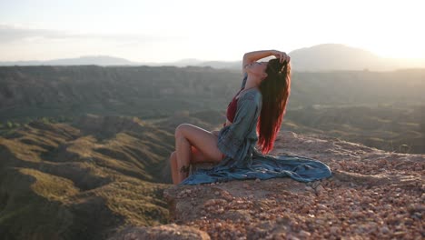 a woman with red hair sits on a cliff edge overlooking a vast desert landscape at sunset