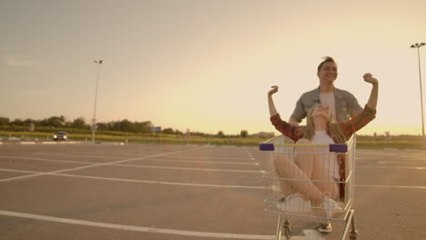 Side-view-of-a-young-man-and-woman-having-fun-outdoors-on-shopping-trolleys.-Multiethnic-young-people-racing-on-shopping-carts.-On-the-parking-zone-with-their