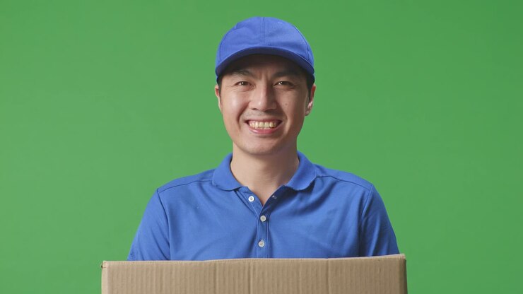 Primo piano di un corriere maschio asiatico in uniforme blu che sorride mentre consegna un pacco su sfondo green screen in studio.