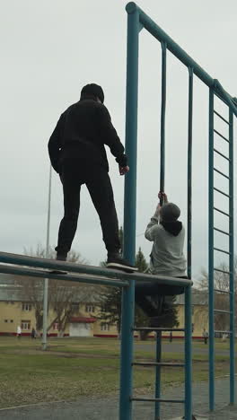 a young boy dressed in a black outfit carefully walks on an iron pole while another person descends the iron rail and walks away, meanwhile, a boy in a gray sweater climbs the iron structure