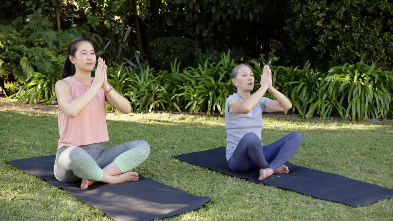 Practicing yoga outdoors, Asian grandmother and granddaughter meditating on yoga mats in garden