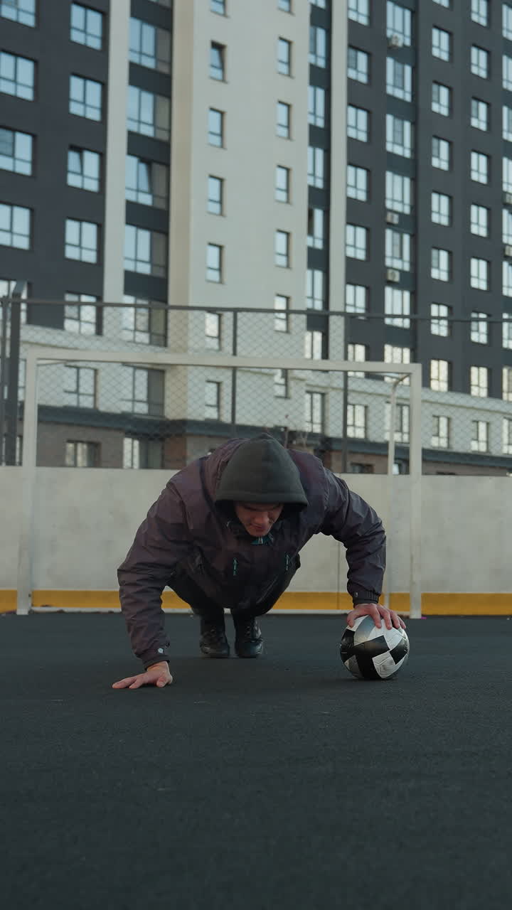 Man performing push ups alternating hand placement on soccer ball, showcasing strength, balance, and fitness in outdoor sport arena with goal post and modern residential buildings