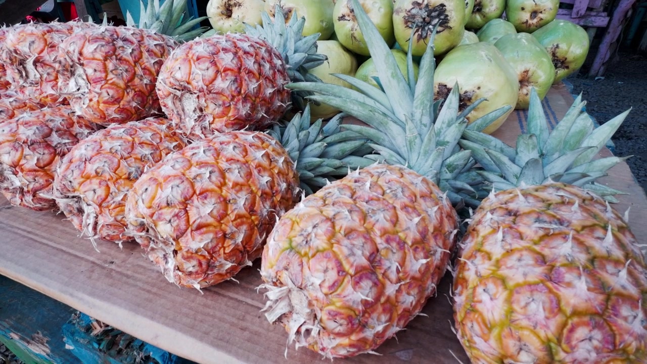 Row of fresh pineapples and pile of green coconuts on display at local fruit market