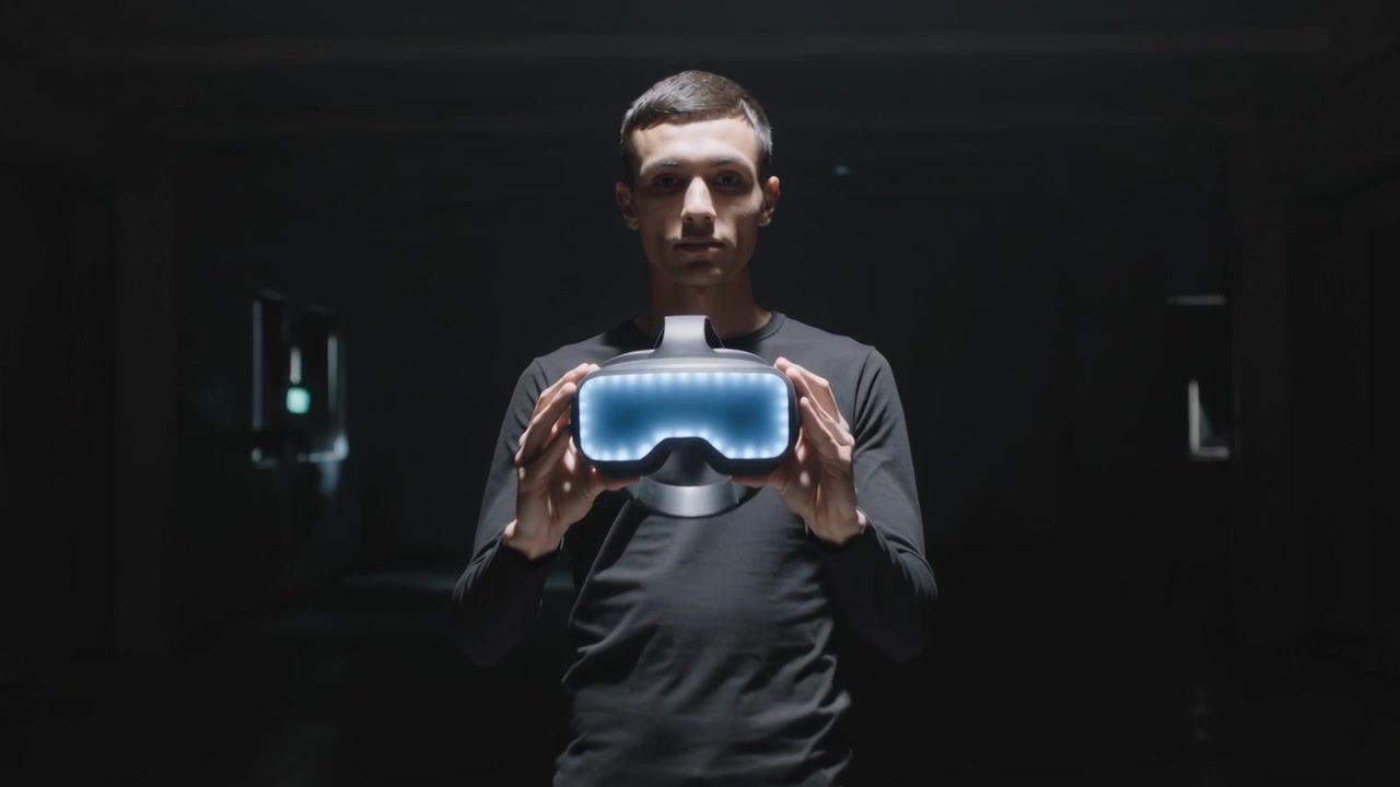Camera zooming, man wearing dark top holding VR headset with blue faceplate showing under spotlight